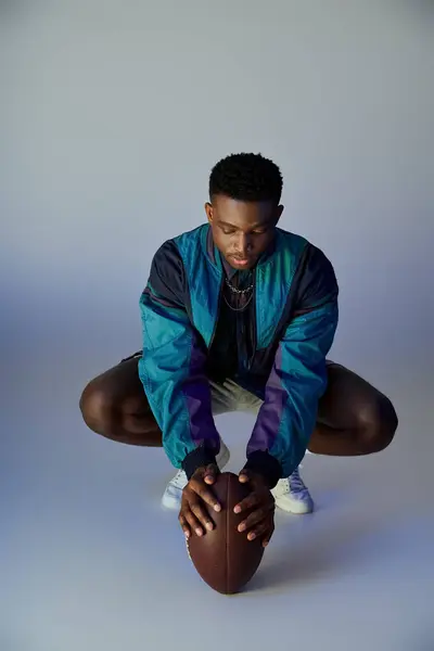 Un joven afroamericano elegante agachándose con un balón de fútbol en sus manos. - foto de stock