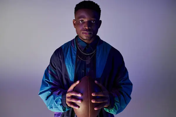 Schöner afroamerikanischer Mann in der Hand eines Fußballs vor einem leuchtend blauen Hintergrund. — Stockfoto
