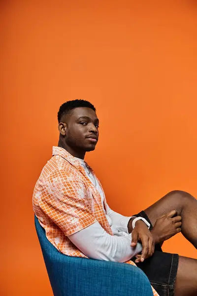 Young African American man in fashionable attire, sitting on chair against vibrant orange background. — Stock Photo