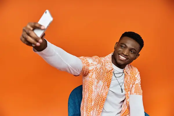 Handsome African American man in trendy attire taking a selfie against a bright orange backdrop. — Stock Photo