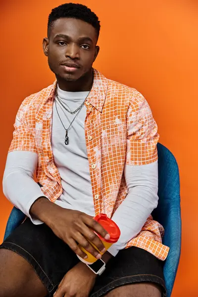African American man in orange shirt looks stylish sitting on blue chair. — Stock Photo