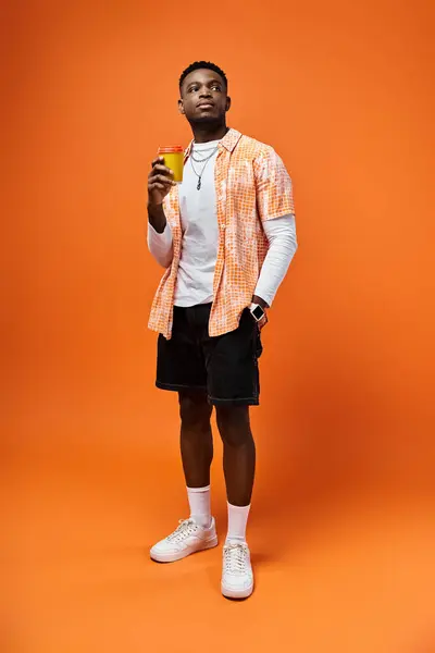 Beau Afro-Américain en tenue tendance tenant un verre devant un fond orange vif. — Photo de stock