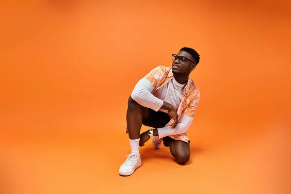 Fashionable African American man crouching on vibrant orange background. — Stock Photo