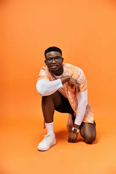 Fashionable young African American man crouching in stylish glasses on bright orange background. — Stock Photo