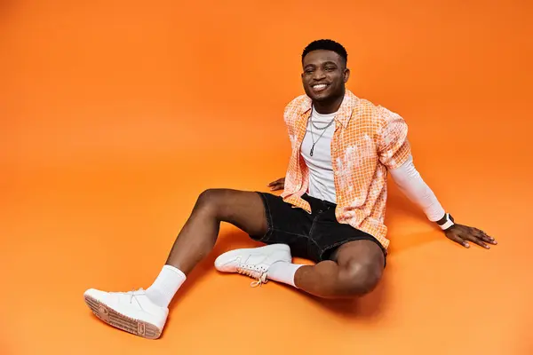Fashionable young African American man in stylish attire sitting on bold orange backdrop. — Stock Photo