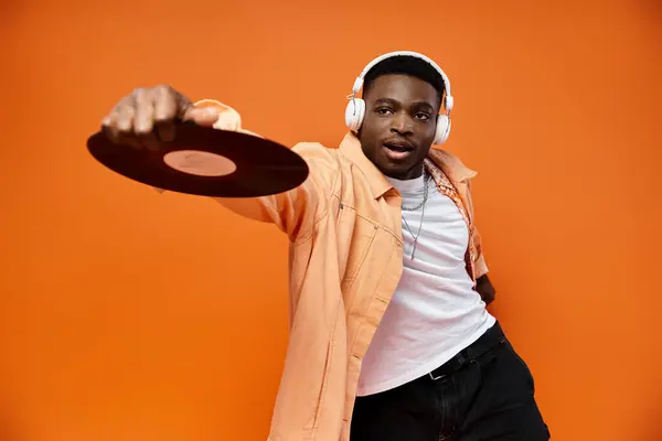 Stylish black man with headphones holding a record on orange background. — Stock Photo