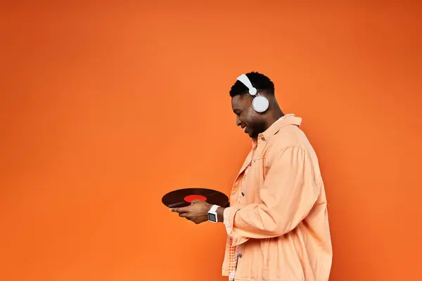 Handsome African American man in headphones holds record on orange backdrop. — Stock Photo