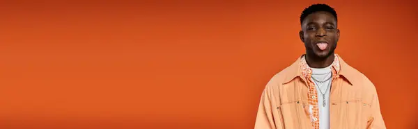 Young African American man in stylish attire sticks out his tongue against orange background. — Stock Photo