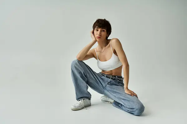 A stylish woman in a white tank top and jeans sitting gracefully on a white background. — Stockfoto