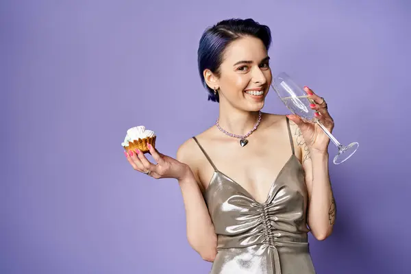 A stylish young woman with short blue hair in a silver dress holds a cupcake and a wine glass in a posed studio shot. — Photo de stock