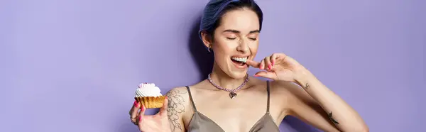 A pretty young woman with short blue hair in a silver party dress joyfully holds and savors a delicious cupcake. — Stockfoto