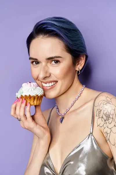 A pretty young woman with short blue hair posing in a silver party dress, holding a cupcake in front of her face. — Stockfoto