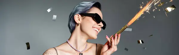 A stylish woman with short blue hair poses in a silver party dress, wearing sunglasses and holding a sparkler. — Photo de stock