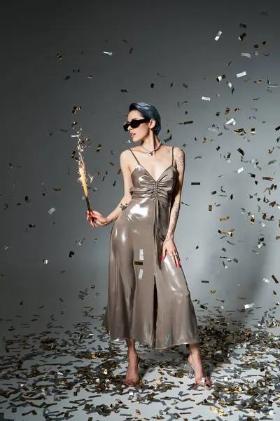 A young woman with short blue hair elegantly poses in a silver dress, holding a sparkler in a mystical studio ambiance. — Foto stock