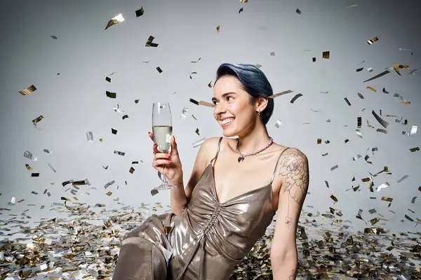 A pretty young woman with short blue hair sits on the ground, elegantly holding a glass of champagne in a silver party dress. — Stock Photo