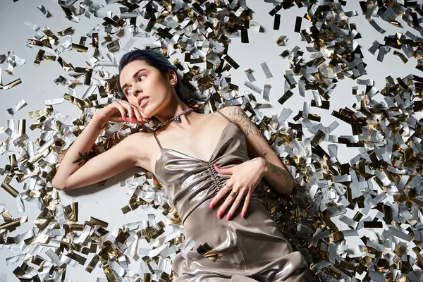 A young woman with blue hair poses elegantly in a silver party dress while laying on a floor of confetti. — Fotografia de Stock