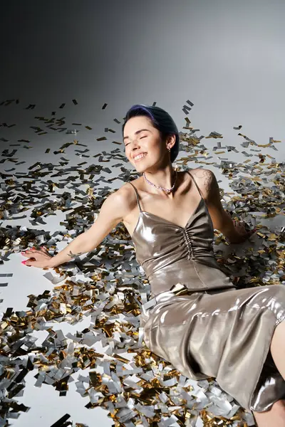 A pretty young woman with short blue hair sitting on a pile of confetti in a shimmering silver party dress. — Fotografia de Stock