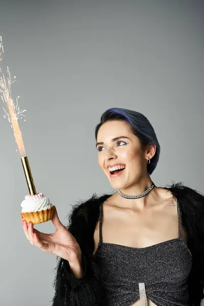 Young woman in stylish attire holding cupcake with lit sparkler in hand, celebrating a special occasion. — стоковое фото