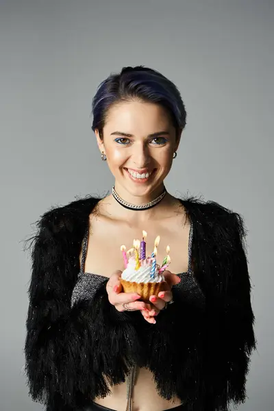Young woman with short dyed hair in stylish attire holds up a cupcake adorned with candles, looking happy and celebratory. — стокове фото