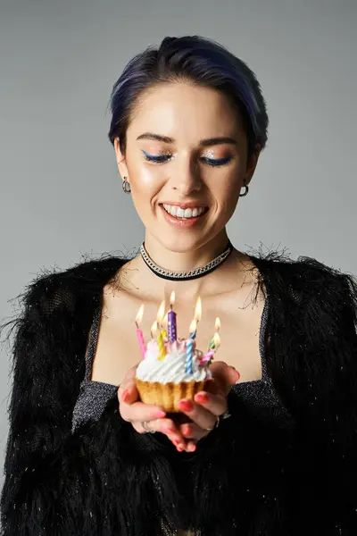 A young woman with short dyed hair holding a festive cupcake with lit candles, dressed elegantly in a studio setting. — Stock Photo