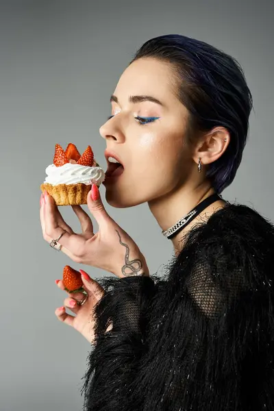 A fashionable young woman with short dyed hair enjoys a cupcake topped with strawberries in a studio setting. — стокове фото