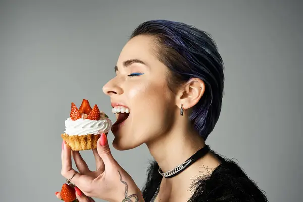 A young woman with short dyed hair smiling while eating a cupcake topped with strawberries. — стокове фото