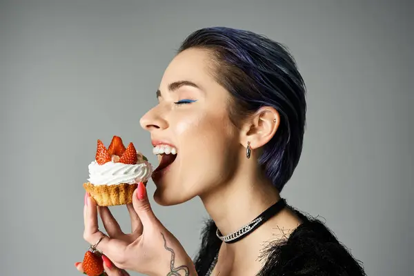 A young woman with short dyed hair smiling while eating a cupcake topped with strawberries. — Stock Photo
