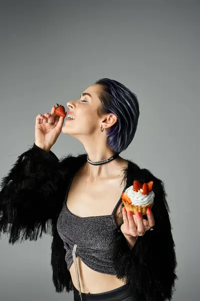 A stylish young woman with short dyed hair, wearing a jacket, enjoys a pastry in a studio setting. — стокове фото