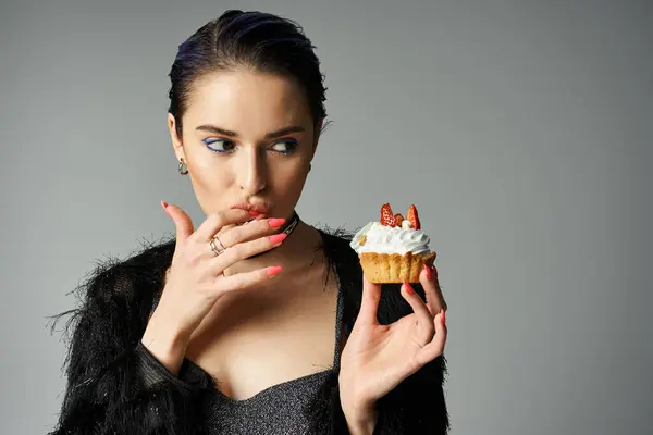 A young woman with short dyed hair poses in stylish attire, holding a delicious cupcake in her hand. - foto de stock