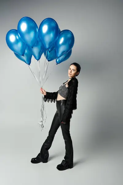 A stylishly dressed young woman with short dyed hair poses gracefully, holding a bunch of blue balloons. — Photo de stock
