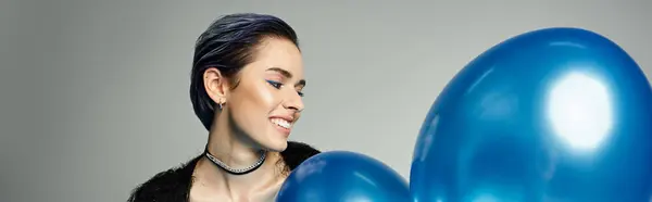 A young woman with short dyed hair holding a bunch of vibrant blue balloons in a stylish studio setting. — Stock Photo