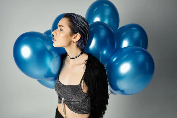 A young woman with short dyed hair balances blue balloons in a stylish studio setting, embodying a whimsical birthday celebration. — Stock Photo