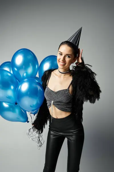 Young woman with short dyed hair in stylish attire smiles, holding a bunch of colorful balloons and wearing a party hat. — Foto stock