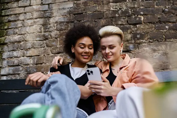 Zwei Frauen, ein vielfältiges und schönes lesbisches Paar, sitzen auf einer Bank in einem Café, in ihr gemeinsames Handy vertieft.. — Stockfoto