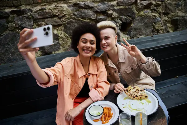 Um casal diversificado de mulheres desfrutando de uma refeição juntas em uma mesa, selfie. — Fotografia de Stock