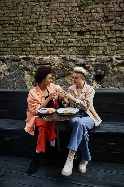 Дві жінки, різноманітна лесбійська пара, насолоджуються їжею разом на лавці в кафе. — стокове фото
