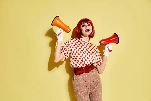 Eine rothaarige Frau mit Pop-Art-Make-up hält zwei leuchtende Megaphone vor gelbem Hintergrund. — Stockfoto