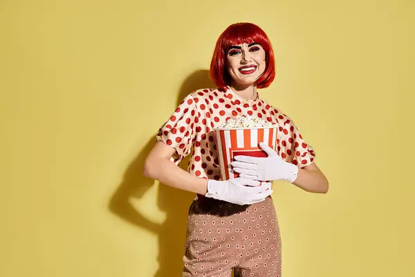 A pretty redhead woman in polka dot blouse with pop art makeup holding a box of popcorn on a yellow background. — Stock Photo