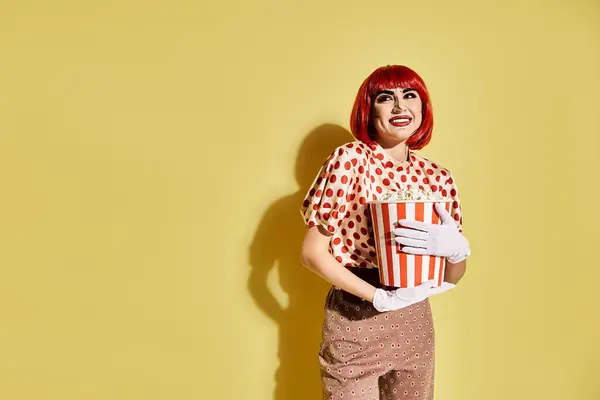 A pretty redhead woman with pop art makeup holds a box of popcorn against a vibrant yellow background. — Stock Photo