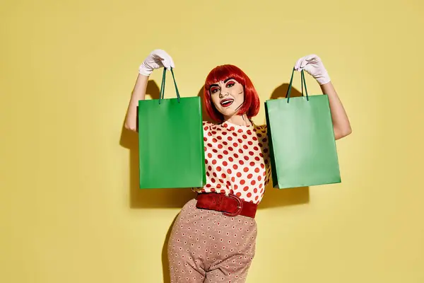 A stylish redhead woman in pop art makeup and polka dot blouse holding shopping bags on a vibrant yellow background. — Stock Photo
