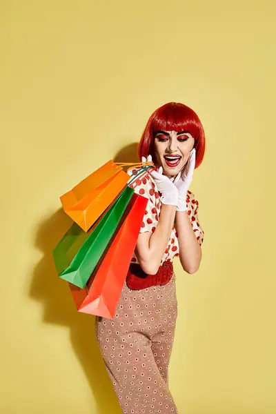 A stylish redhead woman with pop art makeup holding colorful shopping bags against a vibrant yellow backdrop. — Stock Photo