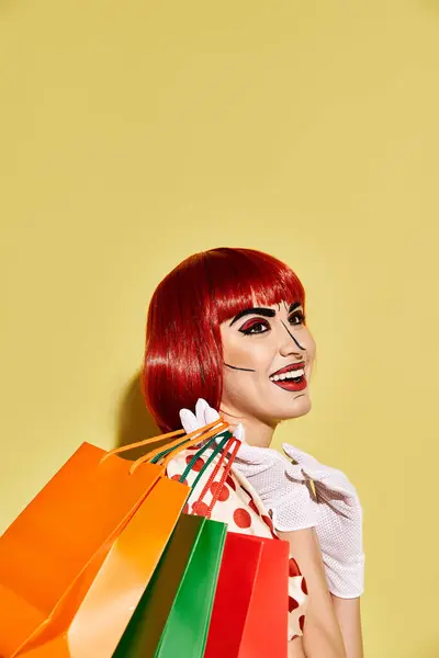 Vibrant redhead woman with pop art makeup and polka dot blouse happily holds colorful shopping bags on a yellow background. — Stock Photo