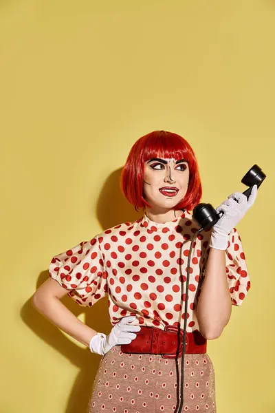 Eine lebhafte rothaarige Frau mit kreativem Pop-Art-Make-up und gepunkteter Bluse hält ein Telefon auf gelbem Hintergrund. — Stockfoto
