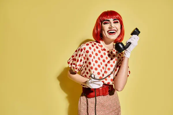 Eine stylische rothaarige Frau mit Pop-Art-Make-up hält ein Telefon vor gelbem Hintergrund. — Stockfoto