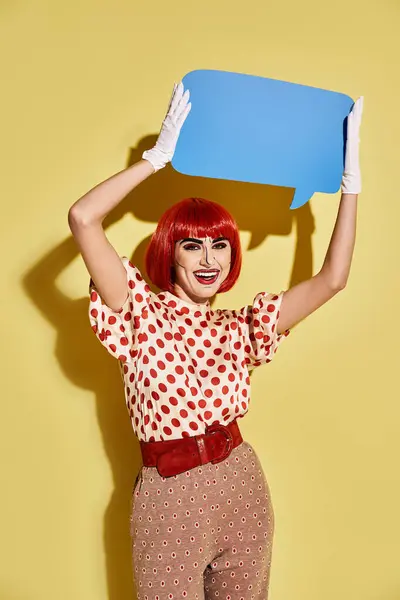 A creative redhead woman in pop art makeup and polka dot blouse holds a vibrant blue speech bubble on a yellow background. — Stock Photo