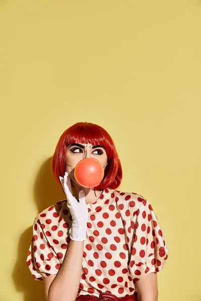 A pretty redhead woman with pop up makeup, dressed in polka dot blouse, blows a bubble on a yellow background. — Stock Photo