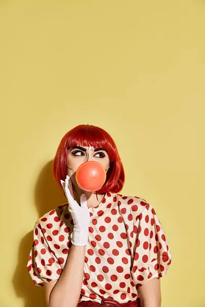 Una bella donna rossa con trucco pop-up, vestita con camicetta a pois, soffia una bolla su uno sfondo giallo. — Foto stock