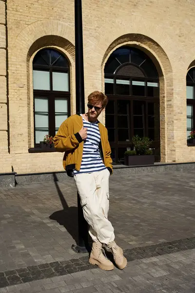 Стильный молодой человек с рыжими волосами уверенно стоит перед кирпичным зданием в городе. — стоковое фото