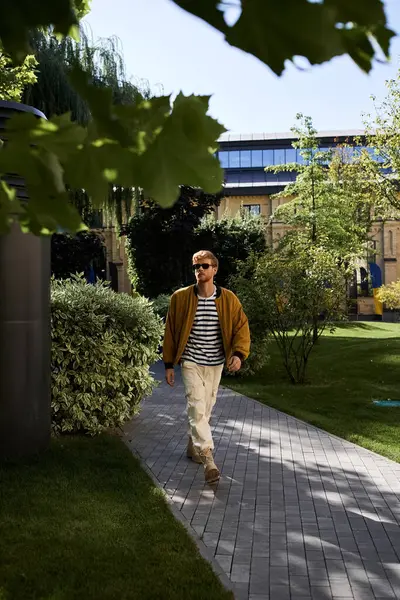 A young red-haired man in debonair attire taking a leisurely walk down a city park sidewalk. — Stock Photo