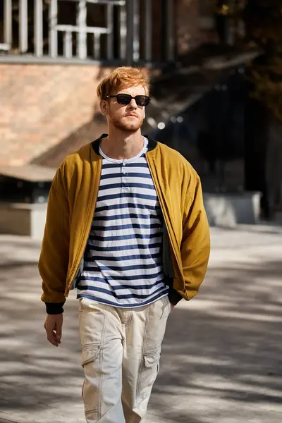 Un giovane uomo dai capelli rossi che cammina fiducioso per una strada della città, indossando occhiali da sole chic. — Foto stock